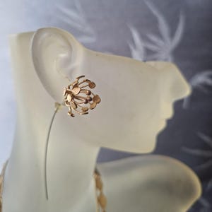May include: A gold-toned stud earring featuring a cluster of small, circular, and spherical elements arranged in a spherical, dandelion-like shape. The earring is attached to a thin, curved metal wire that extends downward from the back of the stud.