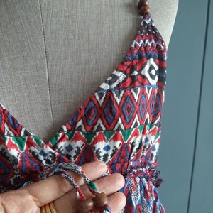 May include: A red, blue, and green patterned sleeveless dress with a V-neckline and a drawstring tie at the waist. The dress has wooden beads on the straps and a fringe trim at the bottom.