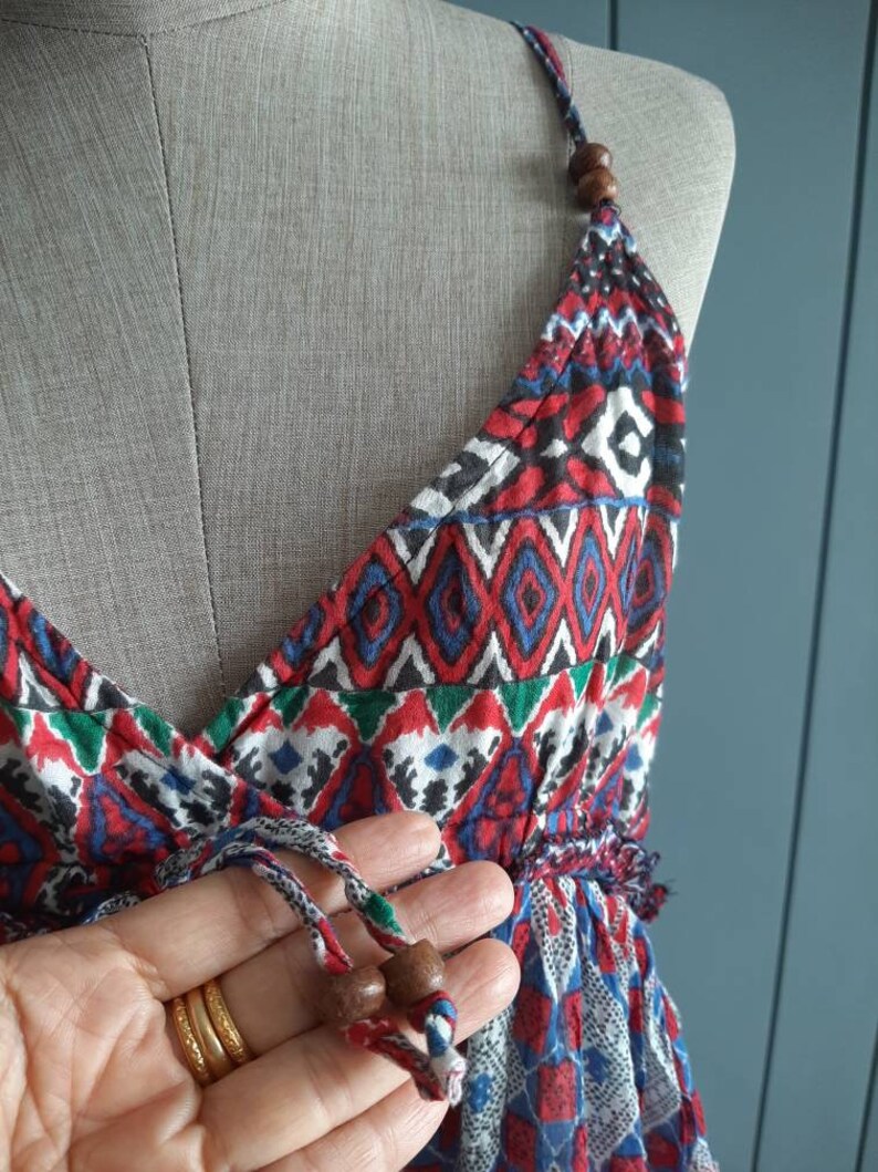 May include: A red, blue, and green patterned sleeveless dress with a V-neckline and a drawstring tie at the waist. The dress has wooden beads on the straps and a fringe trim at the bottom.