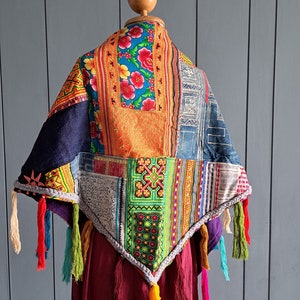 以下が含まれることがあります： 青、赤、黄の花柄、緑とオレンジの幾何学模様の刺繍、藍染めの生地など、様々な布地をパッチワークした三角形のショール。縁はグレーの編み込みトリムとタッセルで仕上げられています。