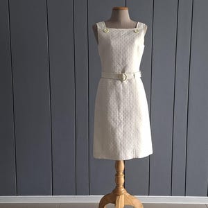 May include: A sleeveless cream-coloured dress featuring a square neckline, two decorative buttons at the shoulders, and a matching belt with a round buckle. The fabric has a subtle, repeating geometric texture throughout the entire garment.