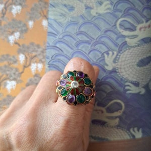 Könnte beinhalten: Ein goldener Ring mit einem großen, bunten Edelstein-Cluster. Der Ring hat einen zentralen weißen Stein, der von grünen, violetten und roten Steinen umgeben ist. Die Steine sind in einer goldenen Fassung gefasst.