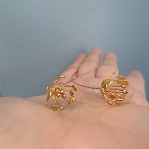 May include: A pair of gold-toned earrings featuring a delicate, multi-strand floral design with small, rounded tips. The earrings are connected by a thin, silver-colored wire and include clear plastic earring backs for secure attachment.