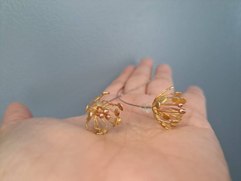 May include: A pair of gold-toned earrings featuring a delicate, multi-strand floral design with small, rounded tips. The earrings are connected by a thin, silver-colored wire and include clear plastic earring backs for secure attachment.
