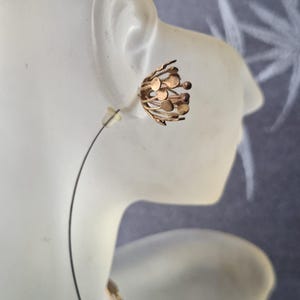May include: A gold-toned earring featuring a spherical, open-work design composed of thin, radiating metal wires topped with small, flat circular discs. The earring is secured with a clear plastic backing and a thin, curved metal wire post.