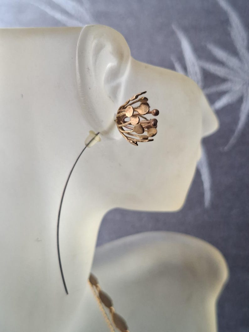 May include: A gold-toned earring featuring a spherical, open-work design composed of thin, radiating metal wires topped with small, flat circular discs. The earring is secured with a clear plastic backing and a thin, curved metal wire post.