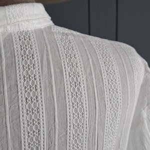 May include: A white long-sleeved shirt featuring vertical stripes of intricate lace detailing alternating with solid fabric panels. The garment has a structured collar and a textured surface, showcasing a repeating geometric pattern throughout the design.