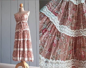 May include: A strapless midi dress in a dusty rose color featuring a floral print with red and green accents. The dress has a tiered skirt design with white lace trim detailing between each tier and a matching fabric tie at the waist.