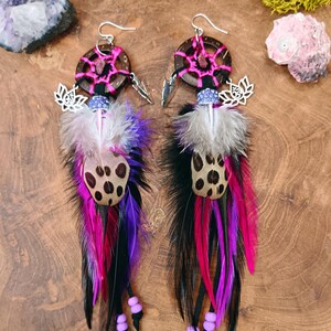 Op de afbeelding: Een paar handgemaakte oorbellen met een cirkelvormige houten basis en een roze webdesign. De oorbellen hebben een combinatie van zwarte, paarse, roze en witte veren, met een gevlekt accentstuk en zilverkleurige bedels.