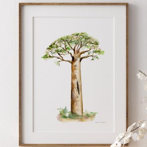 Puede incluir: Una impresión en acuarela enmarcada de un árbol de baobab. El árbol tiene un tronco grueso y texturizado y una copa frondosa verde. La obra de arte se exhibe en una cómoda de madera con una cesta tejida y un jarrón con flores blancas.