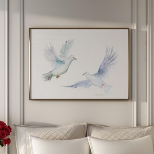 Op de afbeelding: Een wit bed met een wit dekbed en twee witte kussens. Een boeket rode rozen staat op een wit nachtkastje naast het bed. Een ingelijst aquarel schilderij van twee witte duiven in de vlucht hangt aan de muur boven het bed.