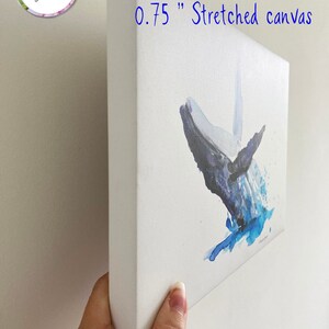 May include: A close-up of a 0.75 inch stretched canvas print of a watercolour painting of a whale jumping out of the water. The canvas is white with a blue and grey whale and blue watercolour splashes.
