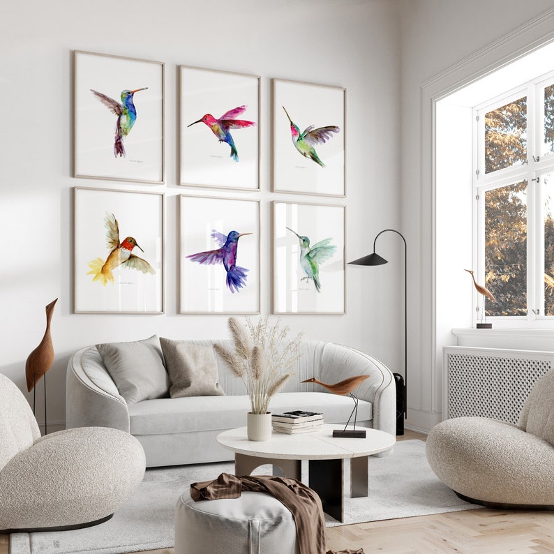 May include: A set of six framed watercolor paintings of hummingbirds in various colors and poses. The paintings are arranged in a grid pattern on a white wall.