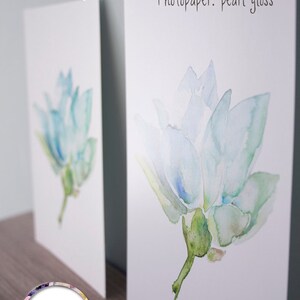 Op de afbeelding: Twee prints van een aquarel schilderij van een lichtblauwe bloem met groene bladeren. De print aan de linkerkant heeft een matte afwerking en de print aan de rechterkant heeft een parelglans afwerking. De tekst "Fine Art: no gloss" staat boven de print aan de linkerkant en de tekst "Photopaper: pearl gloss" staat boven de print aan de rechterkant. De handtekening van de kunstenaar "Michelle Dujardin" staat in een cirkel onderaan de print aan de linkerkant.