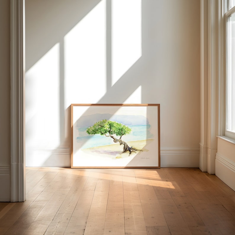 May include: A framed watercolour painting of a tree growing on a beach. The tree is green and has a long, twisted trunk. The beach is white and the water is blue. The painting is in a light wood frame.