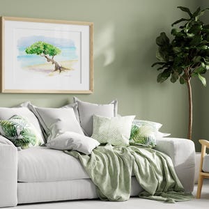 May include: A white sofa with green throw pillows and a green throw blanket. The sofa is in front of a green wall with a framed watercolour painting of a tree on a beach. A green potted plant is in the corner of the room.