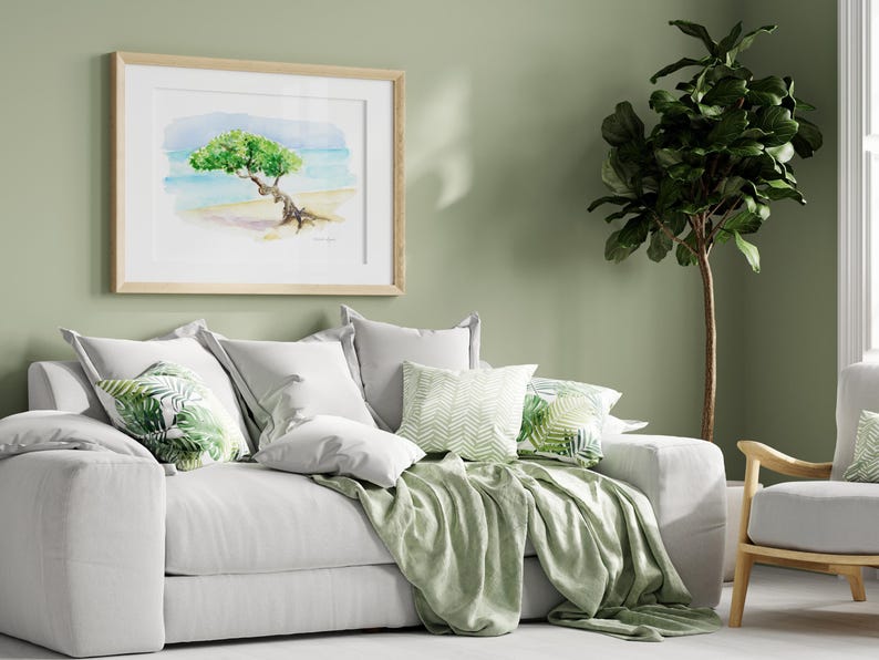 May include: A white sofa with green throw pillows and a green throw blanket. The sofa is in front of a green wall with a framed watercolour painting of a tree on a beach. A green potted plant is in the corner of the room.
