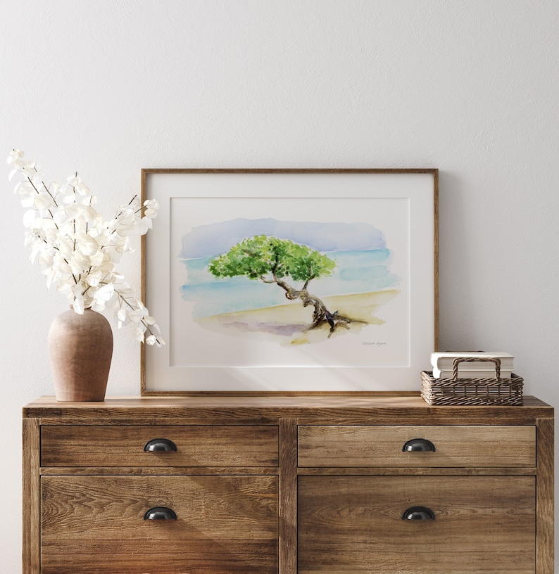 May include: A watercolour painting of a tree bent over by the wind, with the ocean and beach in the background. The painting is framed in a light wood frame and is hanging on a white wall above a wooden dresser.