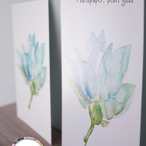 Puede incluir: Dos impresiones de una pintura de acuarela de una flor azul claro con hojas verdes. La impresi&oacute;n de la izquierda tiene un acabado mate y la impresi&oacute;n de la derecha tiene un acabado brillante perlado. El texto "Fine Art: no gloss" est&aacute; por encima de la impresi&oacute;n de la izquierda y el texto "Photopaper: pearl gloss" est&aacute; por encima de la impresi&oacute;n de la derecha. La firma de la artista "Michelle Dujardin" est&aacute; en un c&iacute;rculo en la parte inferior de la impresi&oacute;n de la izquierda.