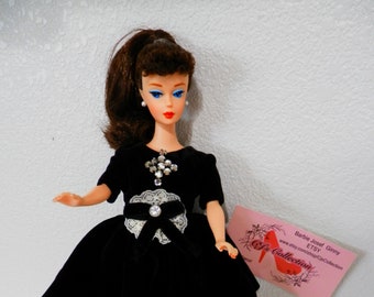May include: A vintage Barbie doll wearing a black velvet dress with a white lace trim and a rhinestone brooch. The doll has brown hair styled in a ponytail and blue eyes. The doll is standing on a white surface with a pink business card that says "Barbie Josef Ginny Etsy" and "www.etsy.com/shop/CipisCollection".