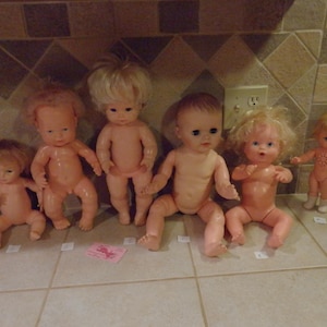 May include: A collection of vintage baby dolls with various hairstyles and poses. The dolls are made of a flesh-toned material and are arranged on a tiled surface. Some dolls are standing, while others are sitting.