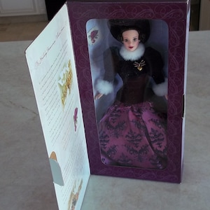 May include: A purple and black doll with a white fur collar and a gold heart pendant. The doll is wearing a long, flowing gown with a black and white pattern. The doll is in a box with a purple background and a white label that says "The Holiday Harmony Collection".