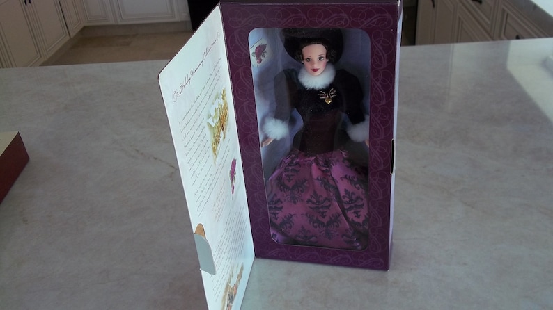 May include: A purple and black doll with a white fur collar and a gold heart pendant. The doll is wearing a long, flowing gown with a black and white pattern. The doll is in a box with a purple background and a white label that says "The Holiday Harmony Collection".