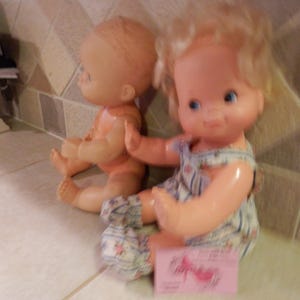 May include: Two vintage baby dolls. One doll is nude, the other doll is wearing a blue and white striped romper with floral accents. The dolls have blonde hair and blue eyes. A pink card is in front of the doll.