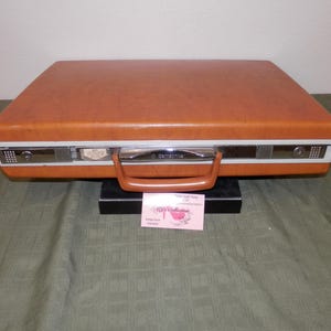 May include: A rectangular Samsonite briefcase with a smooth, burnt orange exterior. The case features a central chrome-colored metal band, a matching orange plastic handle, and two integrated locking mechanisms on the front panel.