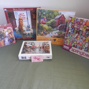 May include: Five different jigsaw puzzles, all 1000 pieces. The puzzles are on a green surface. The puzzles are: Wet Paint, Playing Hookey at the Mall, a Parisian street scene, a scene with a red windmill, and a puzzle with a variety of colorful cans.