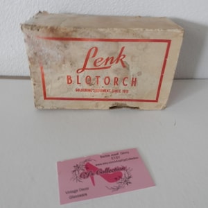 Puede incluir: Una caja de equipos de soldadura vintage Lenk B.L.O.TORCH. La caja es rectangular con un borde y texto rojos. El texto dice "Lenk B.L.O.TORCH Soldering Equipment Since 1910". Una tarjeta de visita rosa está en primer plano.