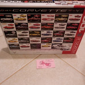 May include: A boxed 1000-piece Corvette puzzle. The box features a grid of various Corvette models in red, yellow, white, and blue. The word "CORVETTE" is prominently displayed. A small pink card is in front of the box.