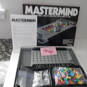 Può includere: Gioco da tavolo Mastermind con una scatola bianca e nera. Il gioco include una plancia di gioco grigia, pioli colorati e istruzioni. La scatola recita "Mastermind" a grandi lettere in grassetto.