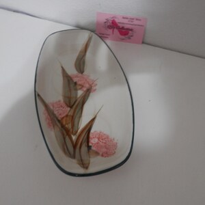 Op de afbeelding: Ovale, witte keramische schaal met een donkerblauwe rand. Het is versierd met een bloemenontwerp met roze bloemen en bruine bladeren. Een klein roze visitekaartje staat op de achtergrond.