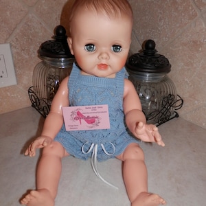 May include: A vintage baby doll with blue eyes and a light blue knit romper. The doll has a pink tag that reads "P Collection" and "Barbie Josef Ginny Etsy". The doll is sitting on a white surface.
