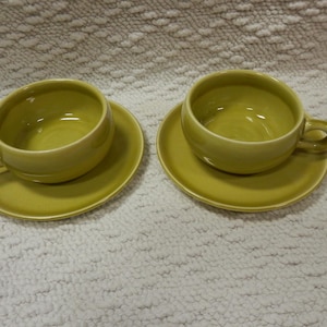 May include: Two matching, light green ceramic teacups and saucers. Each cup has a rounded body, a small handle, and sits on a matching saucer. The set is a solid, muted green color, and the cups and saucers are placed on a textured white surface.