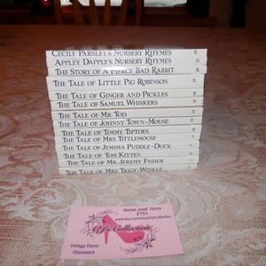 Op de afbeelding: Een stapel kinderboeken met witte covers en titels als "Cecily Parsley's Nursery Rhymes" en "The Tale of Mr. Tod". Een roze visitekaartje met een logo van een schoen met hoge hakken ligt voor de boeken. De boeken liggen op een tafelkleed met patroon.