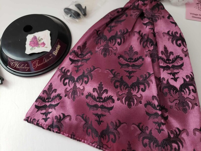 May include: A pink and black patterned fabric skirt with a satin finish. The fabric has a damask design. The skirt is part of a Barbie Holiday Traditions doll outfit.