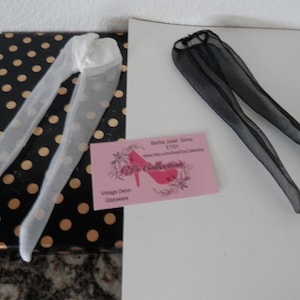 May include: Two pairs of doll-sized tights, one white and one black, on a black and white polka dot background. The white tights are on the left and the black tights are on the right. The black tights are on a white background. The image also includes a business card for "Babe Jewel Story" on Etsy.