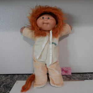 May include: A doll with a round face and brown eyes wearing a cream-colored plush lion costume. The costume features a hood with a thick, orange faux fur mane, a white front panel with white ribbon ties, and an attached orange tail at the base.