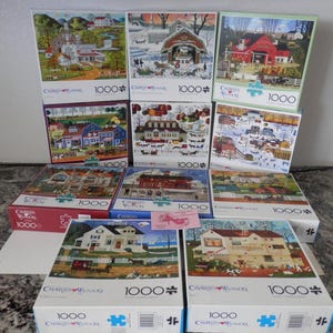May include: A collection of 1000-piece jigsaw puzzles featuring rural scenes. The puzzle boxes display images of houses, barns, and winter landscapes. The puzzles are from the Charles Wysocki collection.