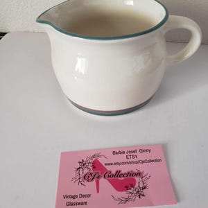 May include: White ceramic creamer with a green and brown rim. The creamer has a rounded body and a handle. The creamer is sitting on a white surface. The image also includes a pink business card with the text "Barbie Josef Ginny ETSY www.etsy.com/shop/CjsCollection CJ's Collection Vintage Decor Glassware".