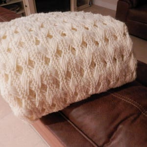 May include: Cream-colored knitted blanket with a diamond pattern. The blanket is folded and resting on a brown leather couch. The knitted texture is visible, with open diamond shapes throughout the design.