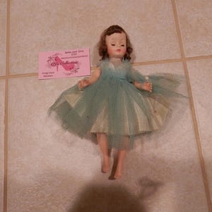 May include: A vintage doll wearing a blue tulle dress with a white underskirt. The doll has brown hair and is posed with arms outstretched. A pink business card is in the background.