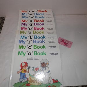 May include: A stack of children's books titled "My 'a' Book" through "My 'x v z' Book". Each book has a colorful cover with illustrations and the text "My First Steps to Reading". The books are stacked on a white surface.