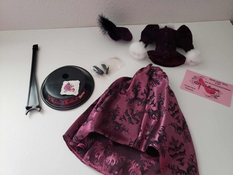 May include: A doll outfit set with a burgundy skirt with a black floral pattern, a burgundy jacket with white trim, a black hat, and a doll stand.