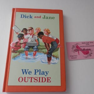 Puede incluir: Libro infantil vintage titulado "Dick and Jane" con una portada naranja. La ilustración de la portada muestra a niños jugando al aire libre en un estanque. El texto "We Play OUTSIDE" se muestra. Una etiqueta rosa con texto es visible.
