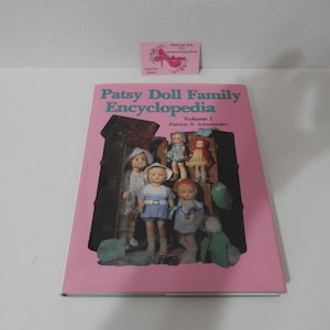 Puede incluir: Un libro rosa titulado "Patsy Doll Family Encyclopedia Volumen 1" con imágenes de muñecas vintage. La portada presenta un baúl marrón oscuro con varias muñecas con diferentes atuendos. Una pequeña tarjeta rosa es visible en el fondo.