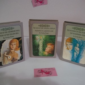 May include: Three vintage paperback books with illustrated covers. The books are titled "The Clue in the Crumbling Wall Mystery of the Tolling Bell", "The Sign of the Twisted Candles Password to Larkspur Lane", and "The Mystery of the Ivory Charm The Whispering Statue". All three books are by Carolyn Keene.