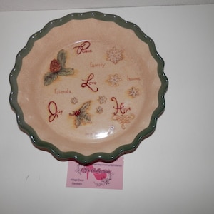 May include: A decorative ceramic pie plate with a scalloped edge. The plate features painted holiday-themed imagery, including snowflakes, holly, and the words "Peace," "Family," "Love," "Friends," "Joy," "Home," and "Hope."